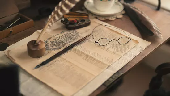 Table avec tasse et soucoupe de porcelaine à côté de la première page d'un journal historique avec des stylos et des lunettes de lecture d'époque sur le dessus.